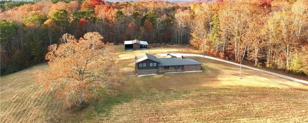 Aerial view of a rural homestead in Upstate South Carolina with a brick home, outbuilding, gravel driveway, and pastures edged by autumn woods